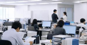Why Japanese Workers Are Judged on Presence, Not Performance Japanese office workers at their desks while one employee stands apart having a conversation, illustrating the tension between visible and invisible work in Japanese workplace culture