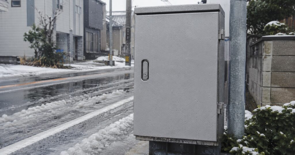 A weathered steel utility box on a Japanese street partially covered in melting snow, muted tones, no visible text or branding.